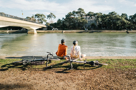 Zwei Radfahrerinnen mit Helmen sitzen am Ufer und blicken auf einen Fluss. Zwei Fahrräder liegen daneben. Im Hintergrund eine Brücke und Bäume.